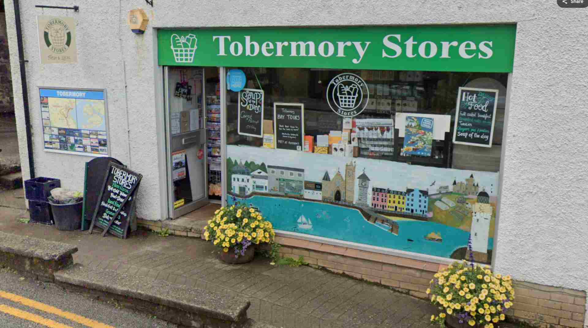 tobermoy stores view from road green signage, shop window and baskets of yellow flowers